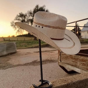 May include: A beige cowboy hat with a brown band and gold trim is displayed on a black metal stand. The hat has a wide brim and a classic western design. The stand is made of twisted metal and has a horseshoe-shaped base.
