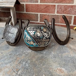 May include: A decorative metal display featuring three dark brown horseshoes welded together. The center horseshoe holds an oval belt buckle with turquoise accents, floral engravings, and the text "I AM COLOSSIANS 3:23". A square silver buckle is attached to the left.