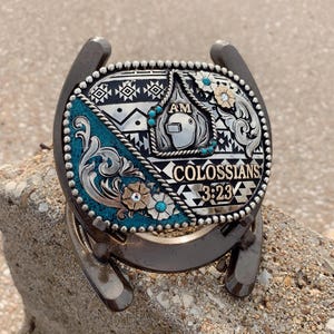 May include: A silver-colored metal belt buckle with a turquoise and silver design, featuring the text "COLOSSIANS 3:23". The buckle is set on a horseshoe base and includes floral and geometric patterns.