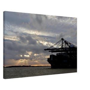 May include: A canvas print depicting a cargo ship silhouetted against a cloudy sky at a port. The dark ship is near a large crane, with the sun's rays breaking through the clouds and reflecting on the water.