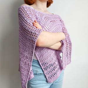 May include: A light purple crochet top featuring an openwork pattern with a wide neckline and short, loose sleeves. The garment has a textured, airy construction with a scalloped hemline and side slits, paired with light blue denim jeans.