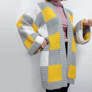 May include: A long-sleeved, open-front cardigan featuring a crochet checkerboard pattern in alternating blocks of bright yellow, white, and light gray. The garment has a ribbed trim along the front opening and wide, loose-fitting sleeves.