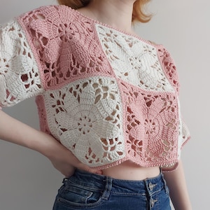Puede incluir: Un top corto de manga corta confeccionado con paneles de ganchillo cuadrados alternos en rosa suave y blanco crema. Cada cuadrado presenta un motivo floral detallado con costuras caladas, creando un patrón geométrico texturizado en la prenda.
