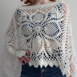 May include: An off-white, cream-colored crochet poncho featuring an intricate, openwork pineapple motif pattern. The garment has a wide, boat-style neckline and a scalloped hemline, showcasing detailed needlework with a delicate, airy texture.