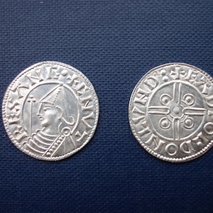 May include: Two silver coins with detailed engravings. One coin features a profile of a figure wearing a hat, surrounded by text. The other coin has a cross design with circular patterns and text around the edge. The coins are on a dark blue surface.