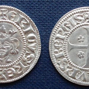 May include: Two silver medieval coins. One coin features a crowned face within a circle, surrounded by text. The other coin displays a cross design with floral accents, also encircled by text. Both coins have intricate details.