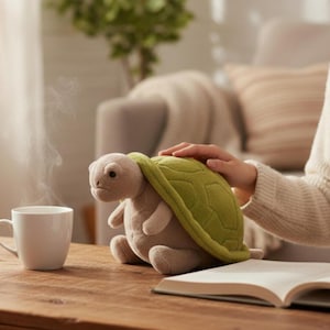 Puede incluir: Un peluche de tortuga con caparazón verde y cuerpo beige está sobre una mesa de madera junto a una taza blanca y un libro abierto. Sale vapor de la taza. La mano de una persona descansa sobre el caparazón de la tortuga.