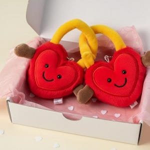 Peut inclure: Deux peluches rouges en forme de cœur avec des visages souriants, des tiges jaunes et des accents marron, présentées dans une boîte blanche avec du papier de soie rose. Les jouets sont conçus avec une expression joyeuse.