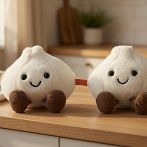 Peut inclure: Deux peluches en forme de bulbes d'ail souriants avec des pieds en velours côtelé marron sont posées sur une surface en bois. Elles sont reliées par un petit cordon orange. Un bol d'ail et une plante en pot sont en arrière-plan.
