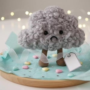 May include: A grey cloud-shaped plush toy with a sad expression, brown legs, and white bandages, presented on a wooden tray with blue tissue paper and pastel sweets. A blank tag hangs from the toy. The background has soft, out-of-focus lights.