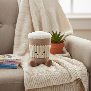 May include: A soft toy coffee cup with a smiling face, a brown body, and a white top, resting on a cream cable-knit blanket. The cup has a ribbed sleeve. A small potted plant and books are in the background.