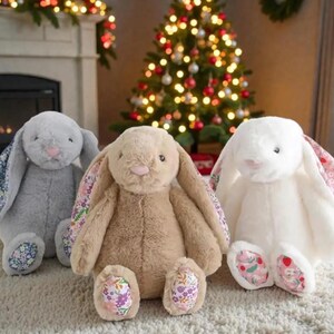 Puede incluir: Tres conejos de peluche en gris, beige y blanco. Cada conejo tiene largas orejas caídas con tela estampada floral en el interior. Los conejos tienen narices rosas y tela estampada floral en los pies. Un árbol de Navidad está al fondo.