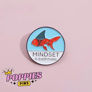 Könnte beinhalten: Runder Emaille-Pin mit einem roten Goldfisch, der eine Haifischflosse trägt, vor hellblauem Hintergrund. Der Pin trägt den weißen Text "MINDSET IS EVERYTHING". Ein Produkt von Poppies Pins.