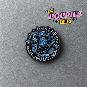 Può includere: Spilla rotonda nera con la scritta "what doesn't kill you mutates and tries again" in bianco. La spilla presenta illustrazioni blu e bianche di microrganismi. La spilla ha la scritta "POPIES PINS" in rosa con una corona d'oro.