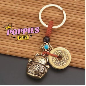 May include: A gold-colored keychain featuring a lucky cat charm, a coin, and decorative beads. The keychain has a red cord and a gold ring. The text "POPIES PINS" is in the upper left corner.