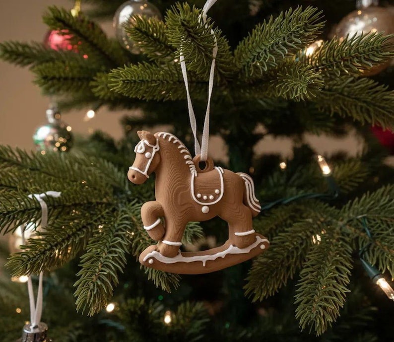 May include: A brown rocking horse ornament with white icing-style detailing on the mane, saddle, and rockers. The horse is suspended by a thin white ribbon, featuring a textured surface that mimics the appearance of a baked gingerbread cookie.