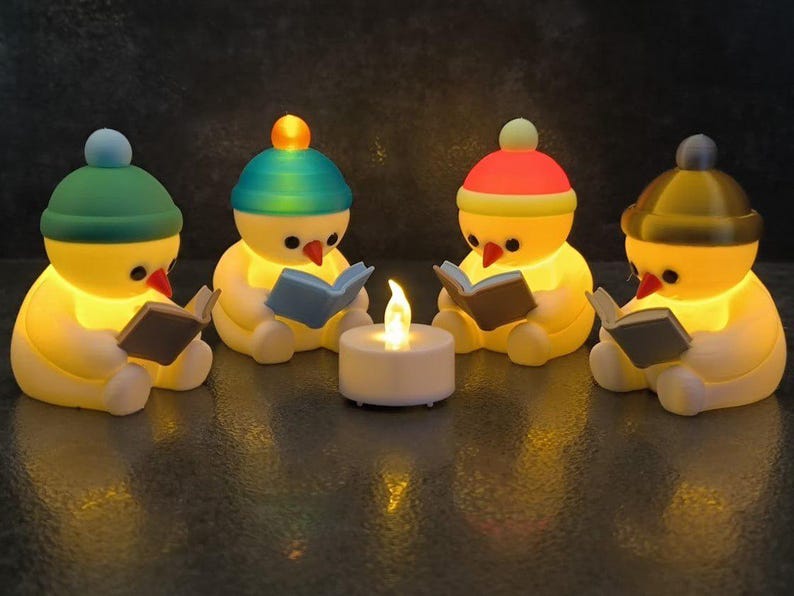 May include: Four yellow, snowman-like figures with orange carrot noses, each wearing a different coloured beanie cap and holding a small book. They are arranged in a circle around a central white, battery-operated tea light with a flickering flame.