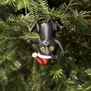 May include: A black cat ornament hanging upside down by its paws. The cat has large yellow eyes and is wearing a red and white Santa hat. The figure is made of a smooth material and features detailed paws with small yellow accents on the toes.