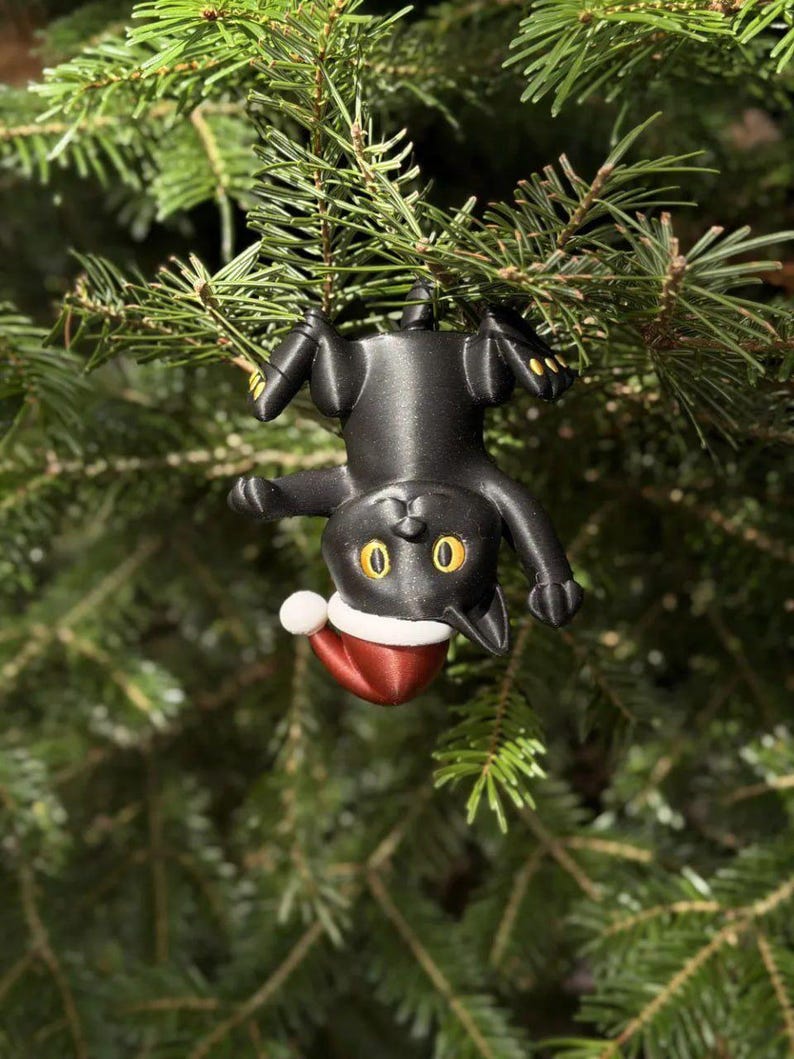 May include: A black cat ornament hanging upside down by its paws. The cat has large yellow eyes and is wearing a red and white Santa hat. The figure is made of a smooth material and features detailed paws with small yellow accents on the toes.