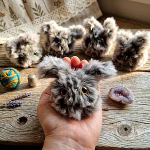 May include: A collection of handmade gray plush bunny toys with orange eyes and fluffy fur. One bunny is held in a hand. Other items include a ball of yarn, a thimble, a sprig of lavender, and a geode on a wooden surface.