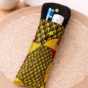 May include: A travel-sized toothbrush holder with a black, yellow, and red patterned fabric exterior. The holder is open, revealing a blue and white toothbrush and a tube of toothpaste. The holder is closed with a yellow button.