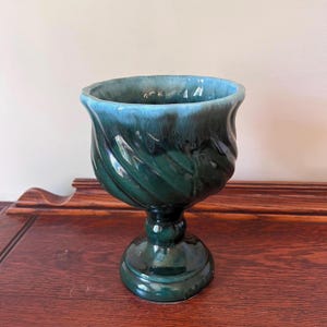 May include: A ceramic chalice with a pedestal base. The chalice is glazed in shades of teal and dark green, with a glossy finish. The body features a spiral design. The rim is a lighter shade of teal. The chalice is displayed on a wooden surface.
