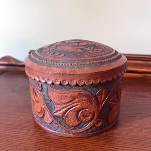 May include: A round, brown leather box with a domed lid. The box features intricate embossed floral and leaf designs. The lid has a detailed embossed pattern. The edges of the box are scalloped.