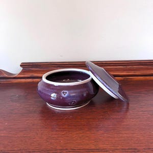 May include: A small, round, purple ceramic jar with a lid, set on a wooden surface. The jar has a white interior and rim, with decorative embossed designs. The lid is slightly ajar, revealing the inside of the jar.
