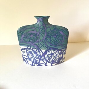 May include: A flat, wide ceramic vase with a narrow neck and a flared rim. The surface features a two-tone design with a teal and purple abstract pattern on the upper section and a white base with a dark blue linear floral motif on the lower part.