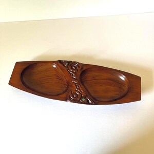 May include: A long, brown wooden serving dish with two oval-shaped compartments. The center section features a carved, textured floral pattern with small, circular green inlays. The wood grain is visible throughout the smooth, polished surface.