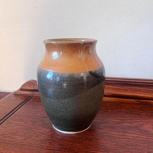 May include: A handcrafted ceramic vase with a two-tone design. The upper portion is a warm, earthy brown, transitioning to a deep, mottled green-blue at the base. The vase has a rounded body and a slightly flared rim, perfect for displaying flowers.
