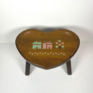 May include: A heart-shaped wooden stool with a dark brown finish. The top surface features painted illustrations of two small houses, a tree with heart-shaped leaves, and a decorative border of alternating light green and pink squares.