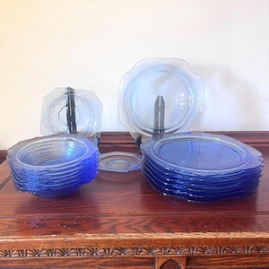 May include: A collection of vintage blue glass dishes, including plates, bowls, and lids. The plates have a square shape with rounded edges. The bowls are stacked, and the lids are displayed on black stands. The dishes are arranged on a dark wooden surface.