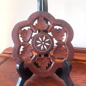 May include: A dark brown, carved wooden trivet with a floral and leaf design. The trivet is supported by a black stand. The center of the trivet features a white starburst design.