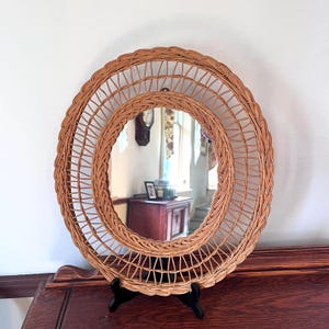 May include: A round, woven wicker mirror with a spiral design. The mirror is supported by a black stand and sits on a wooden surface. The mirror's frame is a light brown color.