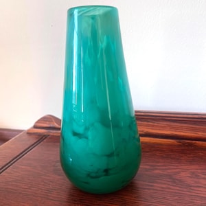 May include: A tall, teal-colored glass vase with a teardrop shape. The vase has a marbled effect, with darker green swirls throughout the teal. The vase is displayed on a dark wooden surface against a white wall.