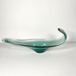 May include: A long, curved glass bowl featuring a deep teal green center that transitions into clear glass edges. The vessel has an elongated, asymmetrical shape with one end sweeping upward into a pointed, curled tip, creating a smooth, fluid form.
