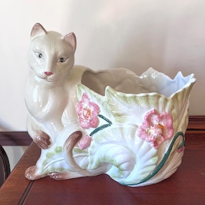 May include: A ceramic cat planter in cream with pink accents. The cat is seated, with a planter shaped like leaves and flowers. The planter has pink flowers and green leaves. The cat has a pink nose and brown ears.
