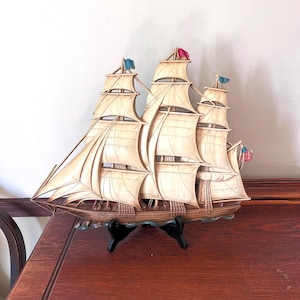 May include: A detailed model of a sailing ship with three masts and multiple sails. The ship is cream-colored with brown accents and features small flags in red, blue, and the American flag. It is displayed on a black stand, showcasing nautical decor.