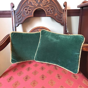 May include: Two rectangular, emerald green velvet throw pillows with gold rope trim. The pillows rest on a red patterned chair. The chair has a dark wood frame with ornate carvings.