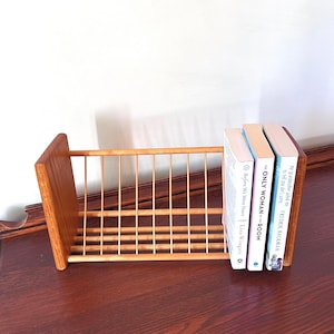 May include: A wooden book rack with a slatted design, holding three books. The books have white and light-colored covers with titles and author names visible. The rack is made of light-colored wood.