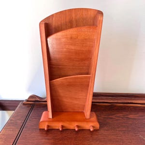 May include: A wooden mail organizer with a warm reddish-brown finish. The organizer has three compartments and a row of four pegs at the base. The design features a curved top and a tapered shape, suitable for home or office use.