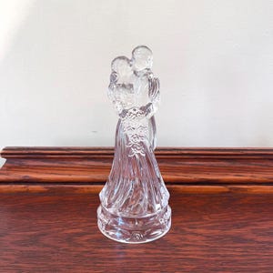 May include: A clear glass figurine of a couple embracing. The sculpture features a man and woman in formal attire, with intricate details in the woman's dress and bouquet. The base is round and detailed.