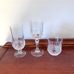 May include: Three clear crystal drinking glasses with diamond-cut patterns. The set includes a champagne flute, a wine glass, and a lowball glass. The glasses are displayed on a dark wooden surface.