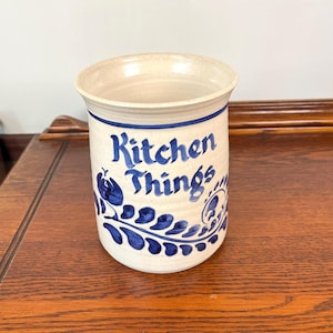 May include: A cylindrical ceramic utensil crock with a cream-colored base and blue hand-painted floral patterns. The text Kitchen Things is written in blue script on the front. The vessel features a wide rim and a smooth, glazed finish.