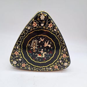 Puede incluir: Una caja de hojalata triangular negra con un borde dorado y un diseño floral colorido. El centro de la caja presenta una escena de una persona montando a caballo a través de un bosque con pájaros volando por encima.
