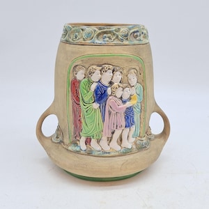 May include: A beige ceramic vase with a relief of figures in colorful robes. The vase has two handles and a green base. The top rim features a decorative pattern with blue and green accents. The figures appear to be singing or holding a scroll.