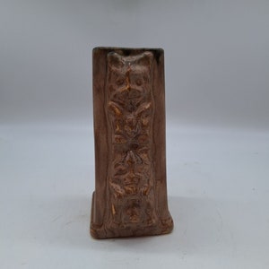 Puede incluir: Un jarrón de cerámica marrón con tres caras talladas. El jarrón es rectangular y tiene una superficie texturizada.