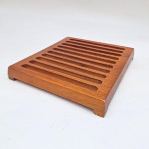 Puede incluir: Una tabla de pan rectangular de madera con una serie de ranuras paralelas. La tabla está hecha de madera de tono medio y tiene pequeños pies. Las ranuras recorren la longitud de la tabla, creando una superficie texturizada.