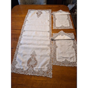 May include: A set of three off-white linen table linens with intricate gray lace trim. The set includes a long table runner and two smaller placemats, all featuring detailed lace patterns and decorative center designs. The linens are displayed on a wooden table.
